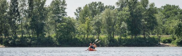 вид издалека на межрасовую активную пару в спасательных жилетах, плавающих на спортивном байдарке с веслами возле реки с зелеными деревьями в летний день, баннер - Фото, изображение