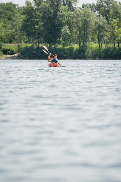 vue de loin de couple sportif multiethnique dans des gilets de sauvetage tenant des pagaies tout en naviguant en kayak sur le lac avec plage pittoresque verte le week-end d'été - Photo, image