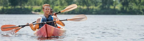 young and sportive redhead man in life vest looking away while holding paddle and sailing on kayak with african american woman on river in summer, banner - Photo, Image