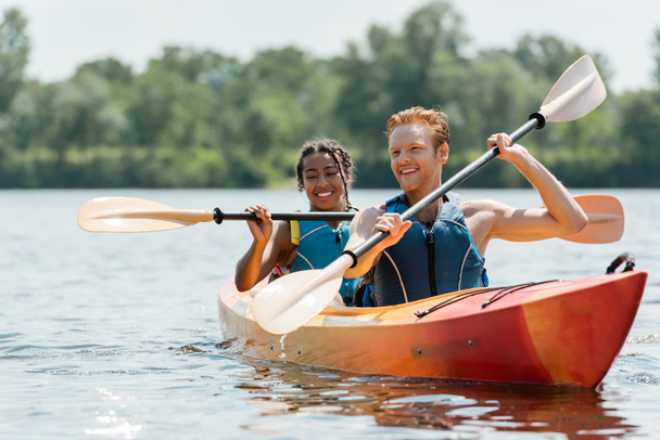 aktywny ruda mężczyzna i uroczy Afryki amerykanka kobieta w kamizelki życia spędzać czas na rzece podczas żeglowania w sportowy kajak na malownicze jezioro na rozmyte tło w lecie - Zdjęcie, obraz