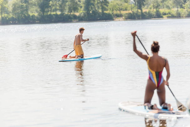 若いスポーツマンがアフリカ系アメリカ人女性の近くの絵のように美しい湖の上でボードに乗って航海するカラフルな水着のパドルボードで夏にぼやけた前景に - 写真・画像