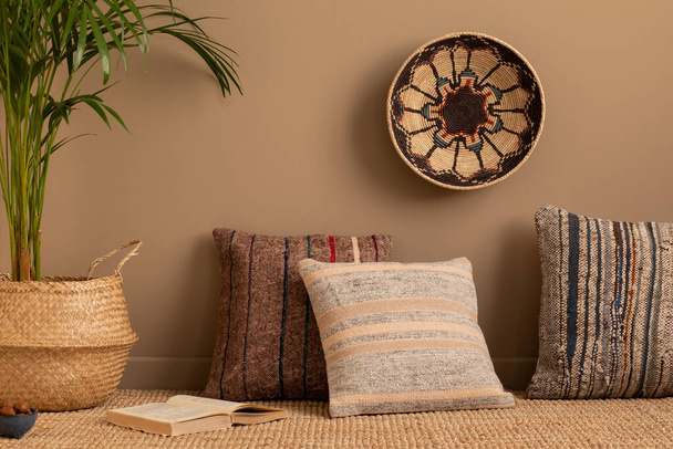 Minimalist composition of ethno living room interior with patterned, colorful pillows, brown wall, braided carpet, round basket on the wall and personal accessories. Home decor. Template.  - Photo, image