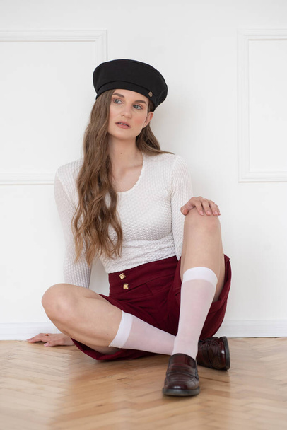 A beautiful young girl is dressed in classic shorts, a white sweatshirt, a beret on her head and wearing loafers in a bright interior - 写真・画像