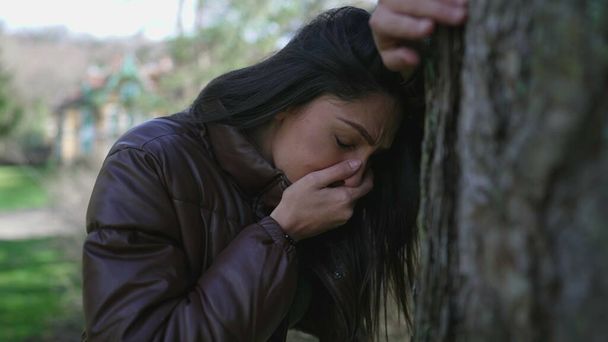 Ağaca yaslanmış acı çeken genç bir kadın çaresiz hissediyor. 20 'li yaşlardaki yetişkin bir kız zor zamanlarda yüzünü utanç ve pişmanlıkla örtüyor. - Fotoğraf, Görsel