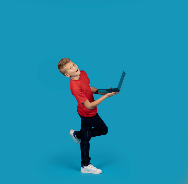 Wesoły chłopiec nastolatek trzyma laptopa i uśmiecha się do kamery, szczęśliwy nastolatek mężczyzna dziecko za pomocą komputera do nauki na odległość i edukacji online, Korzystanie z nowoczesnych technologii, Stojąc na niebieskim tle - Zdjęcie, obraz