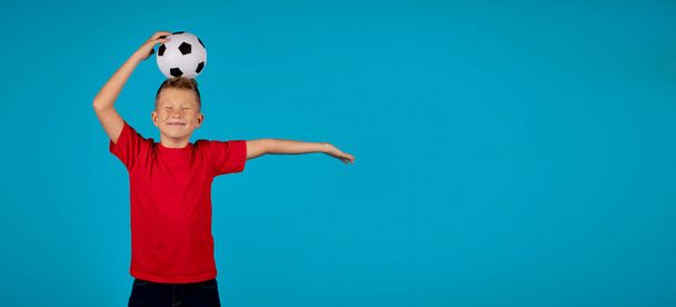 スポーツ広告。スタジオでは、青色の背景に頭の上にサッカーボールでポーズ面白い十代の男の子,目を閉じて立っている男性の子供の間で陽気な,サッカーを楽しんで,コピースペースとパノラマ - 写真・画像