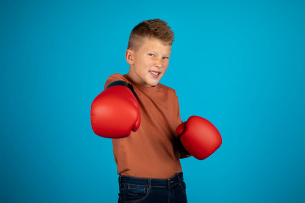 Legrační malý chlapec na sobě boxerské rukavice dělat punč na kameru, veselý předpuberťáci kluk baví při tréninku sportu, Posing izolované přes modré pozadí studia, kopírovat prostor - Fotografie, Obrázek