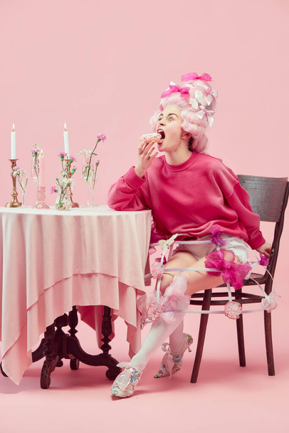 Sweet princess. Portrait hungry princess, queen wearing big pink wig eating donut with exciting face on pink studio background. Comparison of eras, modernity and renaissance, beauty, history concept - Foto, imagen
