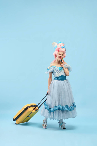 Portrait with smiling princess, queen wearing dress holding suitcase talking via cell phone over blue background. Concept of traveling, comparison of eras, vacation, summer, weekend, relaxation - Photo, Image