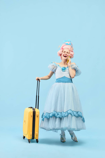 Portrait with happy young princess, queen wearing big pink wig holding suitcase talking via cell phone on blue background. Concept of traveling, comparison of eras, vacation, summer, weekend, emotion - Photo, Image