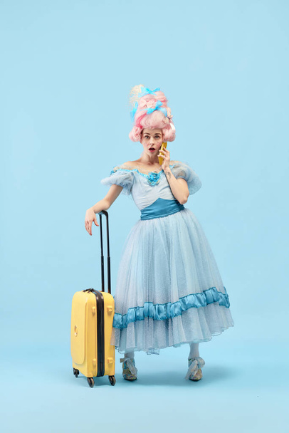 Portrait with surprised young princess, queen wearing big pink wig holding suitcase talking via cell phone on blue background. Concept of traveling, comparison of eras, vacation, summer, emotion - Zdjęcie, obraz