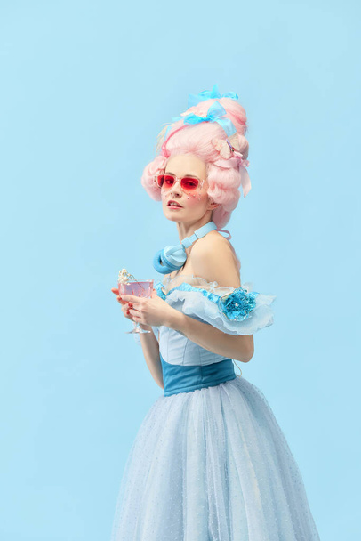 Festa da família real. Retrato de jovem princesa, rainha vestindo vestido elegante, e peruca rosa bebendo coquetel sobre fundo estúdio azul. Conceito de comparação de eras, celebração, relaxante, festa - Foto, Imagem