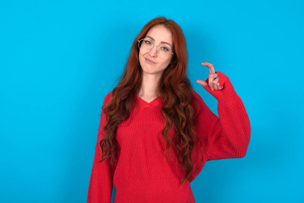 Неймовірна молода жінка в червоному светрі на синьому фоні має маленький знак руки демонструє щось не дуже велике. Концепція мови тіла. - Фото, зображення