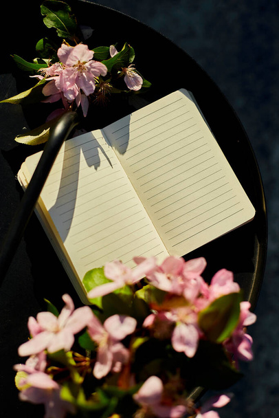 Cuaderno abierto y flores y en la pequeña mesa negra en la terraza en casa en un día soleado, luz dura, espacio de trabajo al aire libre, relajación de verano, notas, ideas, plannin - Foto, Imagen