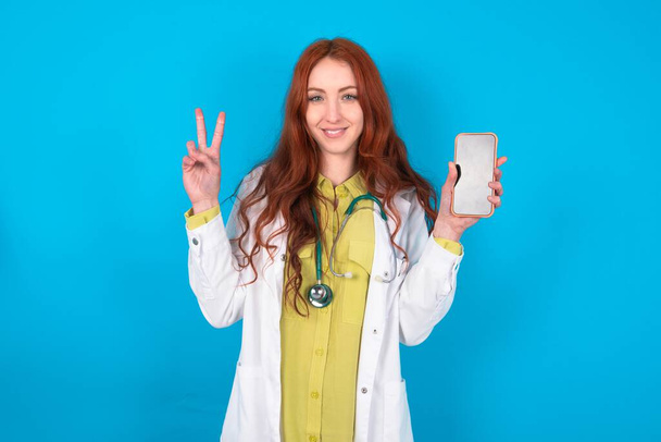 Mladá zrzka běloška lékař žena přes modré pozadí drží moderní zařízení zobrazující V-sign - Fotografie, Obrázek