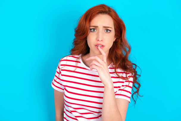 神経質なパズル若い白人赤毛の女性は,青い背景の上にストライプTシャツを着用すると,驚きから口を開き,突然のニュースに反応します. - 写真・画像