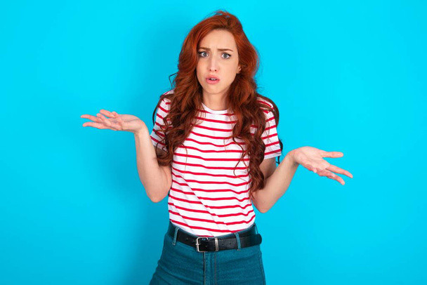 青い背景の上にストライプTシャツを着用したフラストレートヤングアスカシア赤毛の女性は困惑し,躊躇し,恥ずかしがり屋で肩こりは広く開いたまま,何をすべきかわからない. - 写真・画像