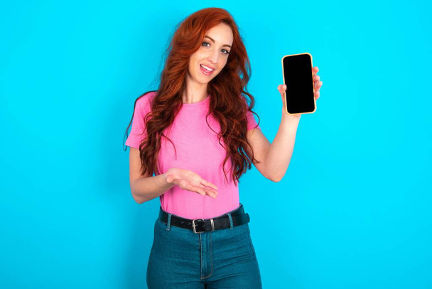 Mladá běloška zrzka žena v růžovém tričku přes modré pozadí s mobilem. prezentuje smartphone. Koncept reklamy. - Fotografie, Obrázek
