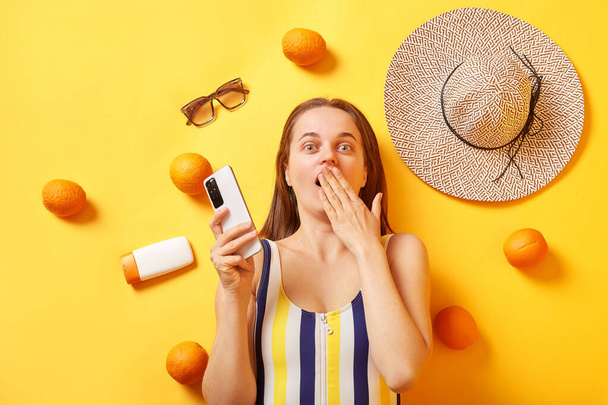 Geschokt verrast vrouw dragen gestreepte zwembroek liggend met sinaasappels zonnebrandcrème op gele achtergrond met behulp van smartphone dekking mond lezen verbaasd nieuws in sociaal netwerk. - Foto, afbeelding