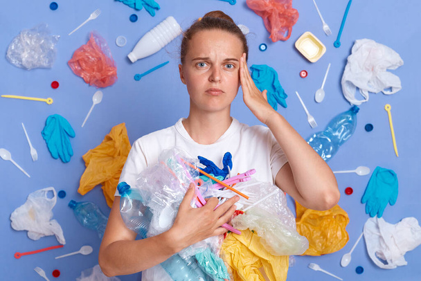 Sad unhappy woman wearing white shirt isolated on blue wall with litter around cleaning territory from rubbish looking at camera with displeased expression. - Foto, Bild