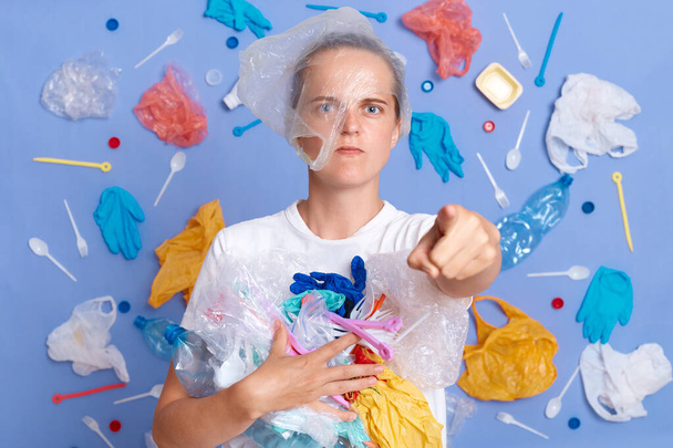 Strict angry serious woman wearing white shirt isolated on blue wall with garbage around cleaning up trash pointing at camera, asking you do not litter. - Foto, immagini