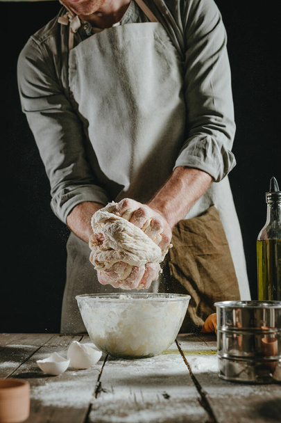 Unrecognizable man in an apron kneading the dough in a glass bowl on a wooden table against black background. Selective focus. - Fotó, kép