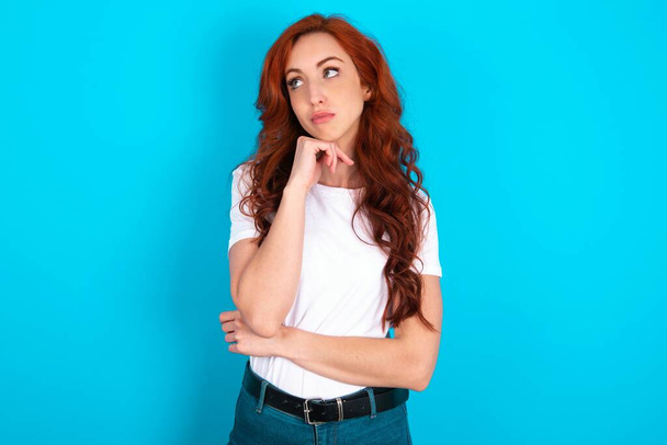 Face expressions and emotions. Thoughtful redhead woman wearing white T-shirt over blue background holding hand under his head, having doubtful look. - Foto, afbeelding