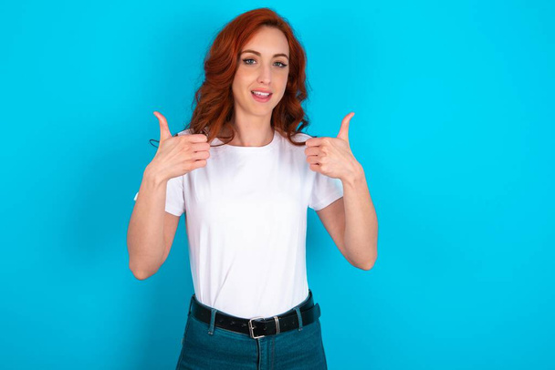 Ottimistica donna rossa che indossa una t-shirt bianca su sfondo blu mostrando i pollici verso l'alto con emozioni positive. Concetto di qualità e raccomandazione. - Foto, immagini