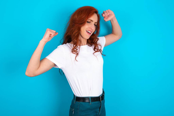 Yes I am winner. Portrait of charming delighted and excited redhead woman wearing white T-shirt over blue background raising up fist in triumph and victory smiling achieving success grinning from delight. - Photo, Image