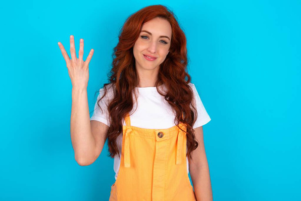 rousse femme portant globalement orange sur fond bleu souriant et regardant amical, montrant numéro quatre ou quatrième avec la main vers l'avant, compte à rebours - Photo, image