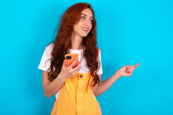 Happy pleased wredhead woman wearing orange overall over blue background man raises palm and holds cellphone uses high speed internet for text messaging or video calls - Фото, зображення