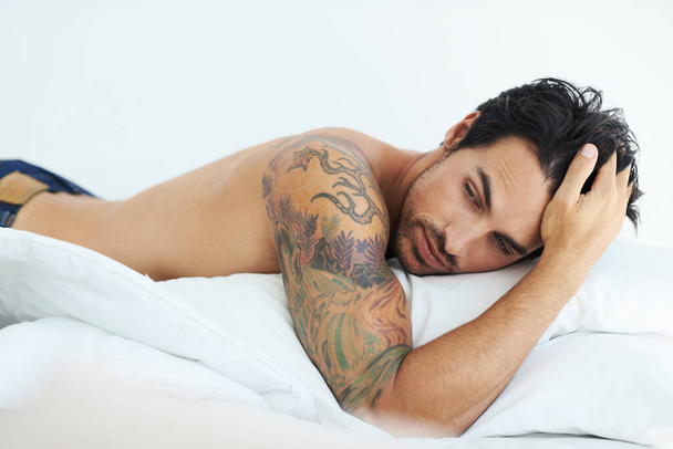 Myślenie, relaks i bez koszulki z seksownym mężczyzną leżącym na łóżku w studio na białym tle. Tatuaż, pomysł i topless z przystojnym młodym modelem pozowanie w sypialni dla zmysłowości lub pragnienia. - Zdjęcie, obraz