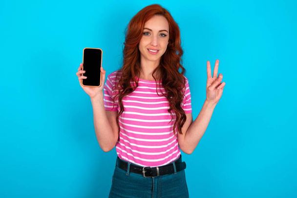 ピンクのストライプTシャツを着た赤毛の女性は,v-signを示す現代的なデバイスを保持する青い背景の上にTシャツ - 写真・画像