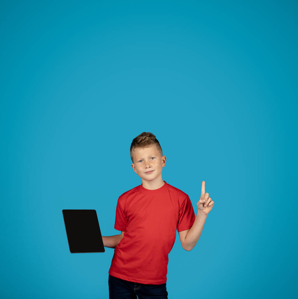 Smiling Male Child Holding Blank Digital Tablet And Pointing Finger Up, Cute Preteen Boy Recommending Study App Or Educational Website While Standing Over Blue Studio Background, Mockup - Photo, Image
