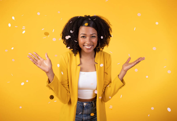 Holiday exuberance. Delighted young black lady ushering in festive cheer, showering confetti over yellow studio background to evoke the spirit of celebration - Zdjęcie, obraz