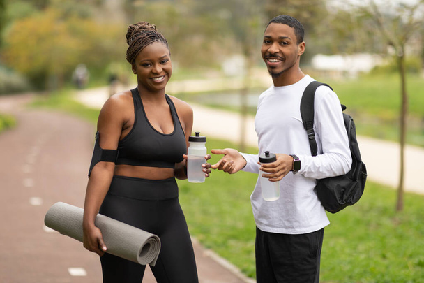 Glad young african american woman and man in sportswear with bottle of water, mat, ready for jogging and fit in park outdoor. Body care, workout in city together, active lifestyle and weight loss - Photo, Image