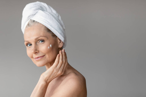 Tratamientos de belleza matutinos para piel envejecida. Feliz anciana en toalla aplicando crema bajo los ojos, mirando y sonriendo a la cámara después de la ducha, espacio para copiar, pancarta - Foto, imagen