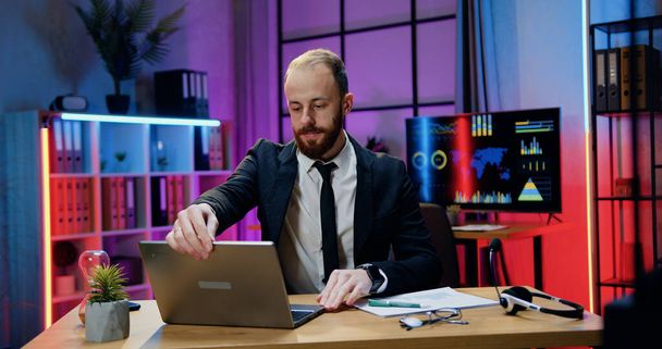 Handsome concentrated industrious responsible skilled bearded businessman sitting at his workplace in evening office and working on computer - 写真・画像