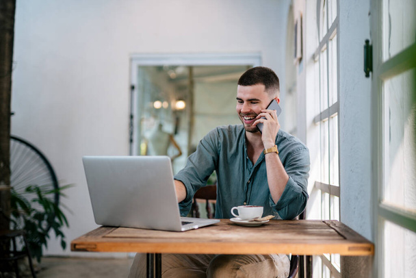 Ein männlicher, gutaussehender Freiberufler lacht, während er telefoniert und an seinem Laptop am Schreibtisch mit seinem Morgenkaffee arbeitet. freiberufliches Konzept. - Foto, Bild