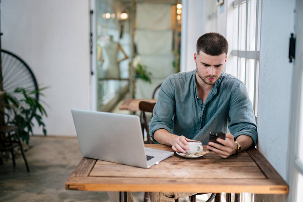 Egy jóképű fiatalember ül kávésbögrével és komolyan ellenőrzi az üzeneteket mobilon laptoppal az asztalon. szabadúszó koncepció. - Fotó, kép