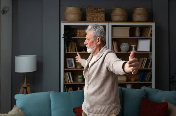 Sporty elderly guy practicing yoga indoors. Senior man doing stretching exercise at home. Active lifestyle and healthcare in any age. Old retired person light fitness training in living room - 写真・画像