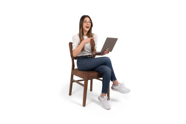 Kobieta wskazująca palcem, podekscytowana biała kobieta trzymająca laptopa kobieta wskazująca palcem. Siedzenie na odizolowanym białym tle, kopiowanie przestrzeni. Polecam stronę internetową, zdjęcie studyjne. Szczęśliwa młoda dziewczyna. - Zdjęcie, obraz