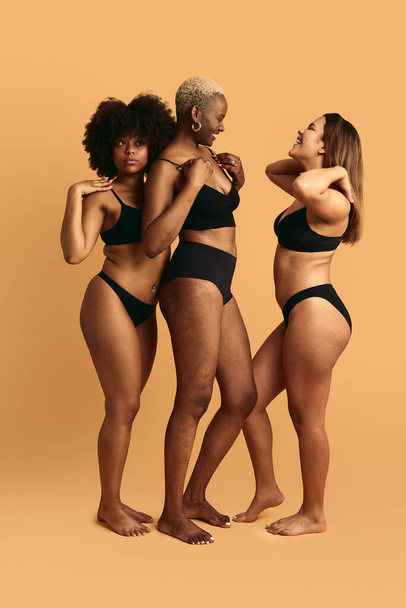 Full body of multiracial women in black lingerie standing in studio touching shoulders fixing hair and adjusting bra against orange background - Photo, Image