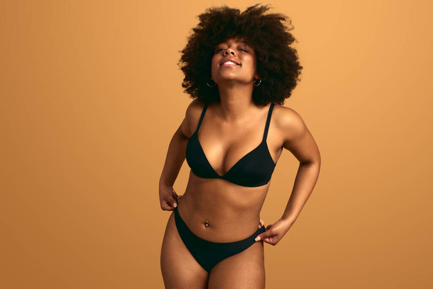 Self esteemed young curvy African American female model in black lingerie with curly dark hair, smiling and looking at camera against beige background in studio - Foto, afbeelding