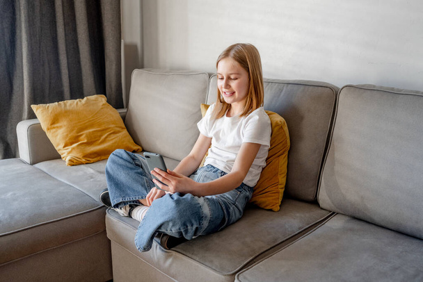 Preteen girl with smartphone sitting on sofa at home. Pretty child kid with modern mobile cellphone on couch - Фото, изображение