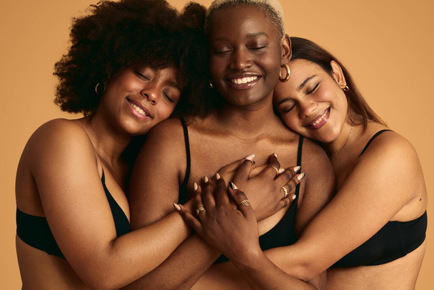 Positive multiracial girlfriends in black tops embracing each other against orange background with eyes closed concept of sisterhood - Fotoğraf, Görsel