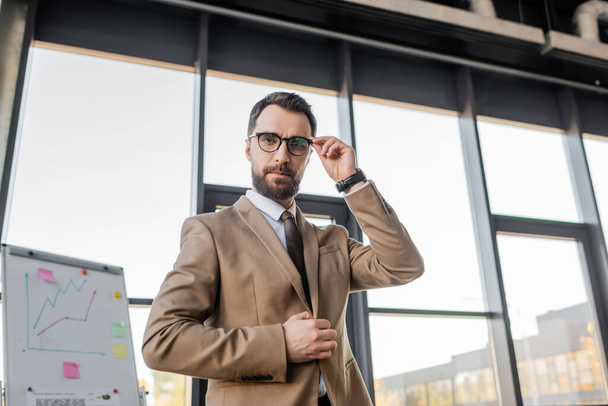 confident manager in stylish formal wear adjusting eyeglasses and looking at camera near flip chart with business planning and analytics on background  - Photo, Image