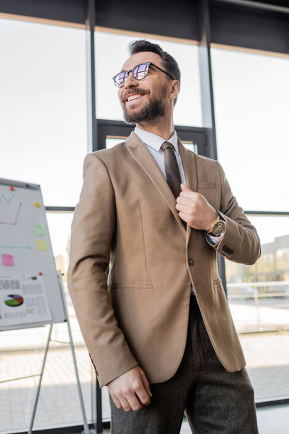 low angle view of successful and happy corporate manager in beige blazer, tie and trendy eyeglasses looking away near flip chart with graphs and business strategy in office  - Photo, Image