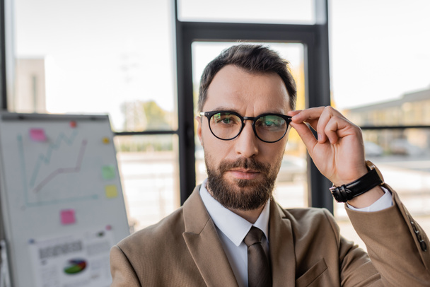 bearded successful businessman in beige blazer and tie adjusting stylish eyeglasses and looking at camera near flip chart with graphs on blurred background  - Photo, Image