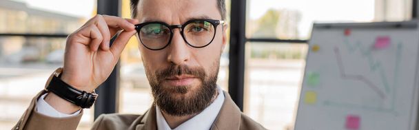 portrait of confident and successful bearded corporate manager in formal wear and wristwatch looking at camera and adjusting trendy eyeglasses in office, banner - Photo, Image